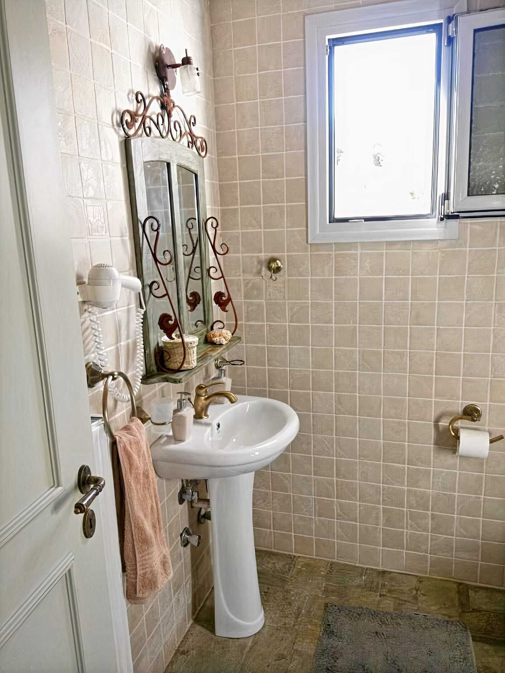 A bathroom with a white sink, a mirror with a decorative frame, a towel hanging on the door, and a window with a view of the outside.