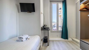 Habitació d'hotel amb un llit, una televisió penjada a la paret, una taula petita amb un safata de recollida de residus, una finestra amb cortina i vista a edificis.