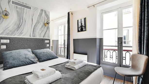 A modern hotel room with a large bed, grey and white bedding, a grey headboard, a chair, and a balcony with a railing. - Cosy Studio for 2P with A/C - Near Montmartre (Paris)