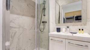A modern bathroom with a glass-enclosed shower, a white marble-tiled floor, a white sink with a mirror above, and a kitchen visible in the reflection. - Cosy Studio - 4P - Palais des Congres (Paris)