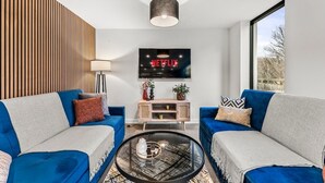 A modern living room with two blue sofas, a glass coffee table, a wooden panel wall, a TV mounted above a wooden cabinet, and a large window.