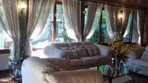 Un vaste salon comprenant un grand canapé beige, un canapé plus petit et une table basse en verre. La pièce est dotée de grandes fenêtres avec des rideaux, d’un plafond en bois et offre une vue sur la verdure environnante.