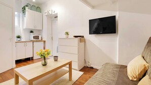A compact living space with a kitchenette, a flat-screen TV mounted on the wall, a wooden coffee table with a vase of flowers, and a sofa with cushions. - Charming studio - 2P- Le Marais (Paris)
