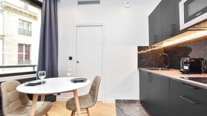 A modern kitchen with dark cabinets, a white countertop, and a coffee machine. There is a round table with two chairs and a wine glass.