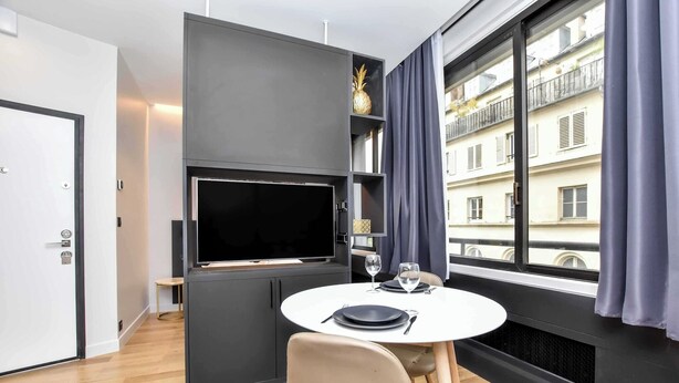 A modern room with a flat-screen TV, a dining table set with plates and wine glasses, and a view of a building through large windows.