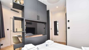 A modern hotel room with a large bed, a flat-screen TV mounted on the wall, a desk with a chair, and a wardrobe with a mirror.