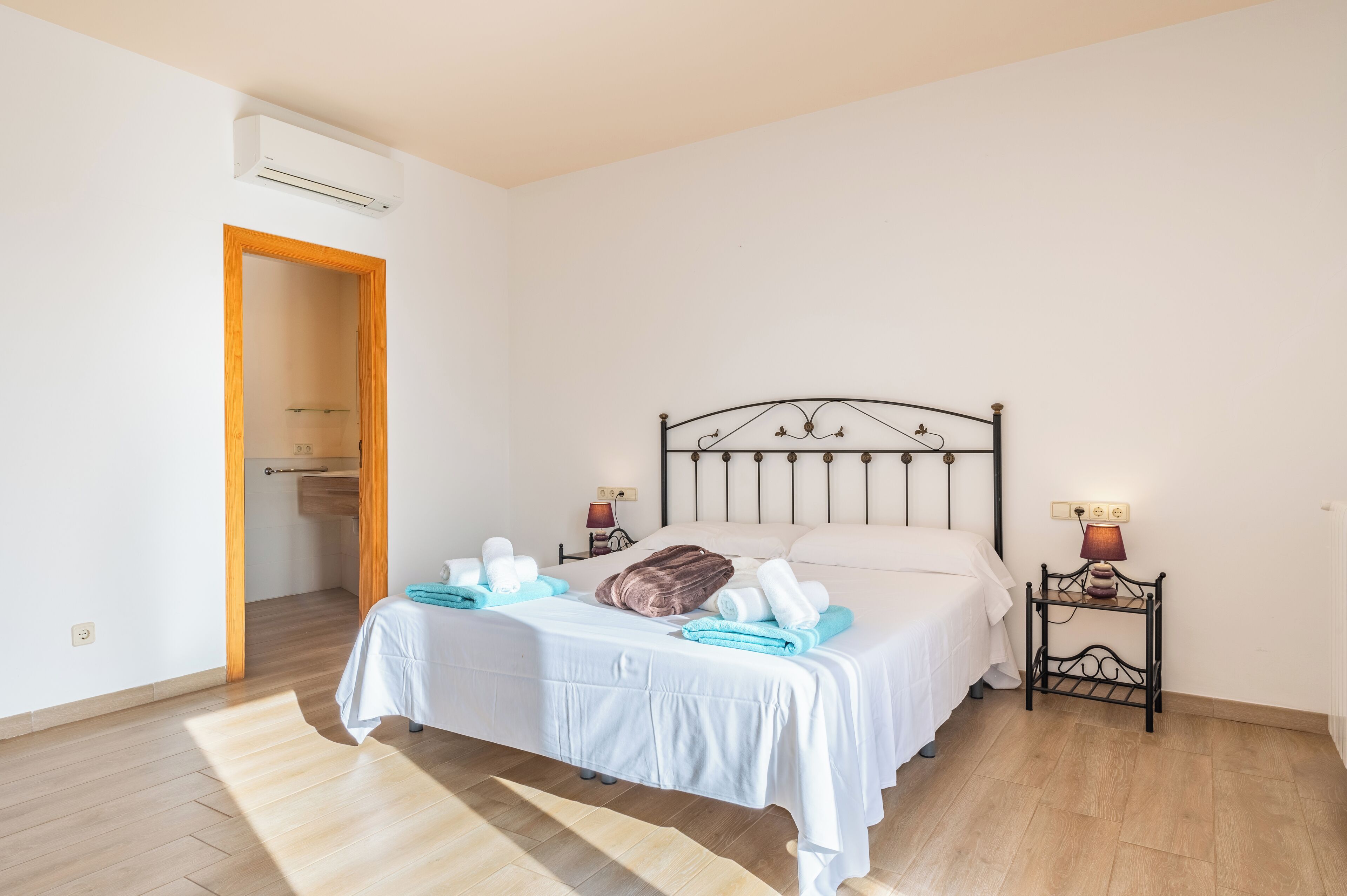 Un lit bien fait, avec du linge de lit blanc et un oreiller marron, une table de chevet avec une lampe, et une porte en bois donnant sur une salle de bain.