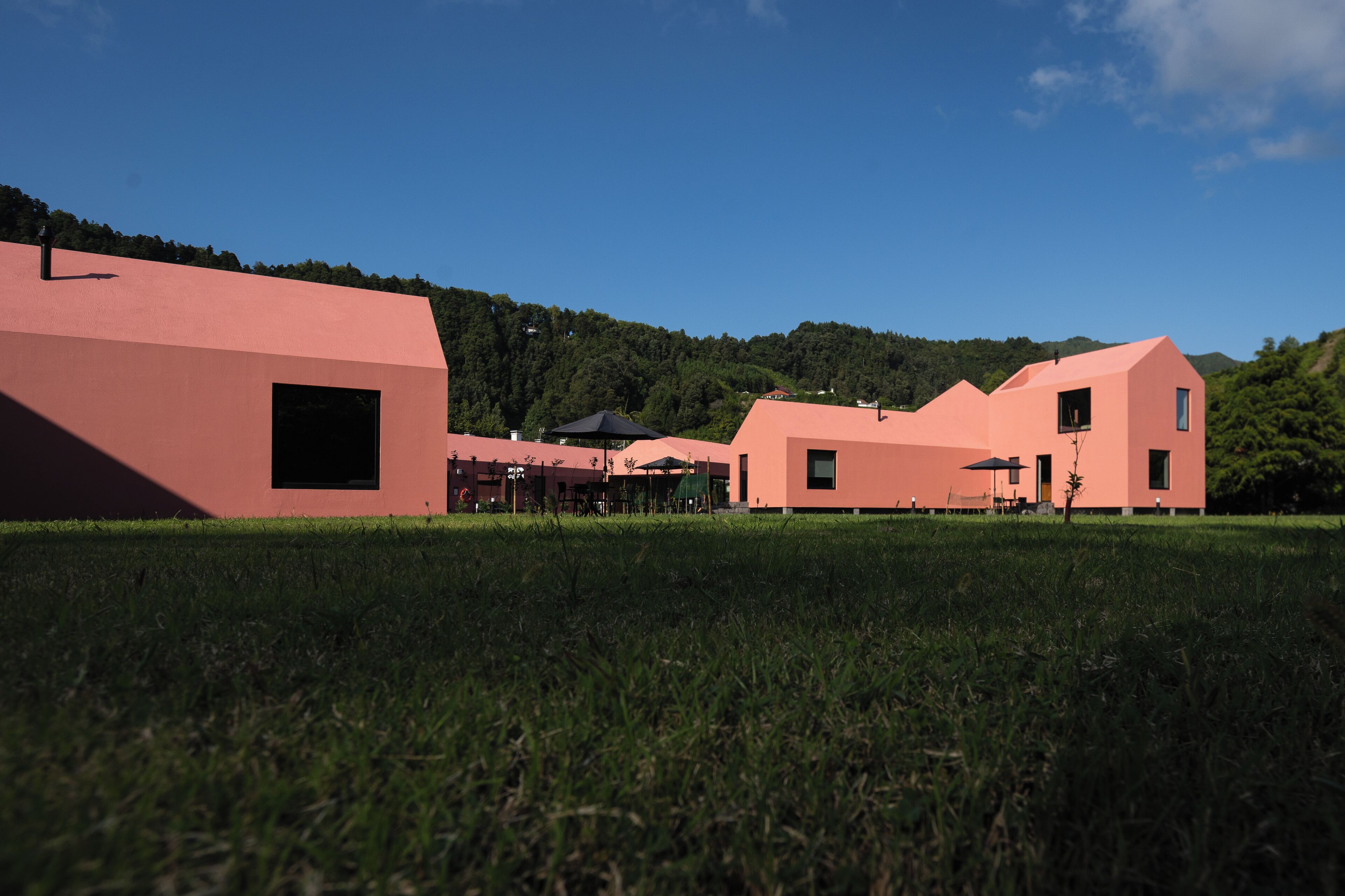 A modern building with a pink facade, surrounded by a grassy field and a forested hill in the background.