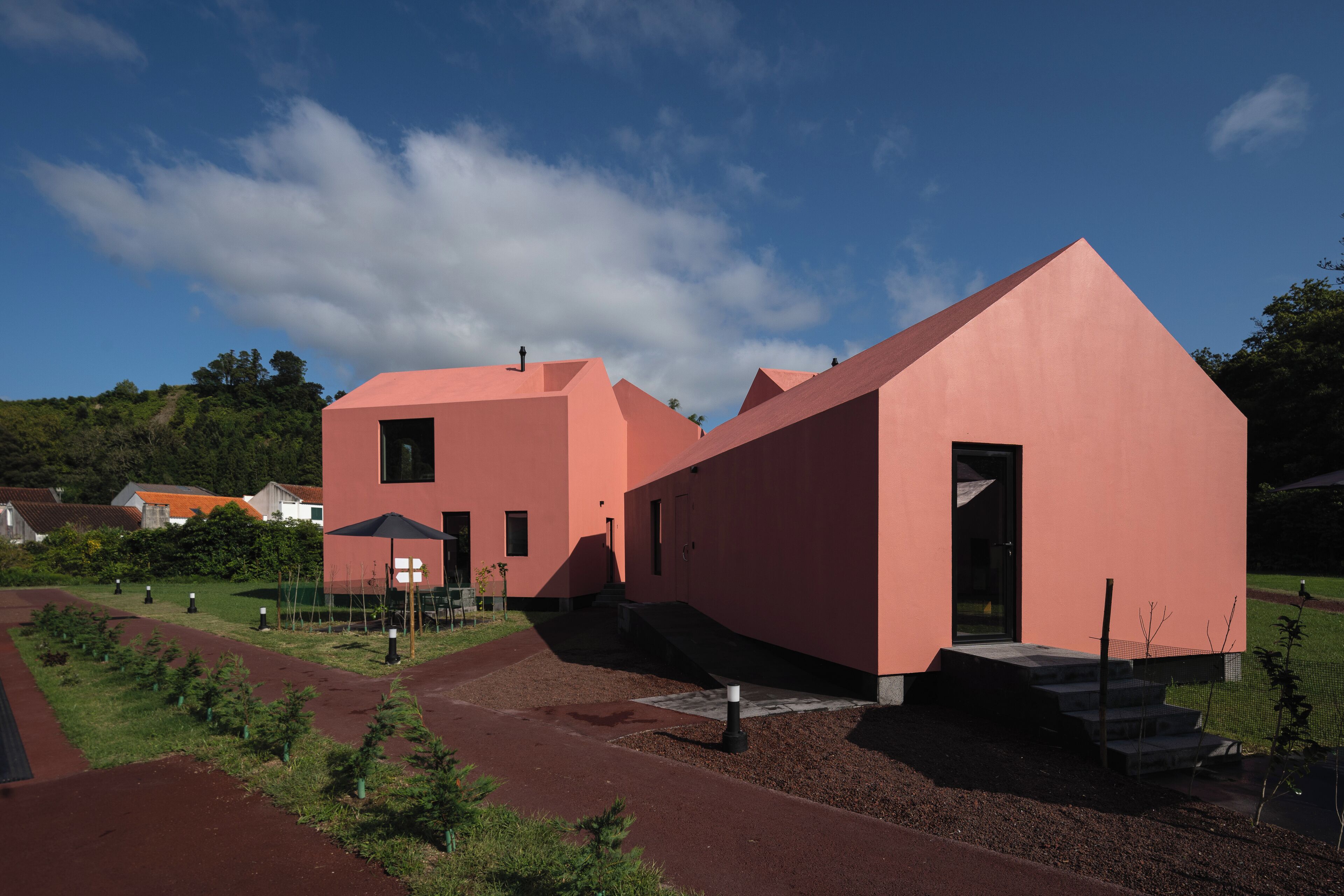 A modern pink building with a sloped roof, surrounded by a landscaped garden with young trees and a gravel path.