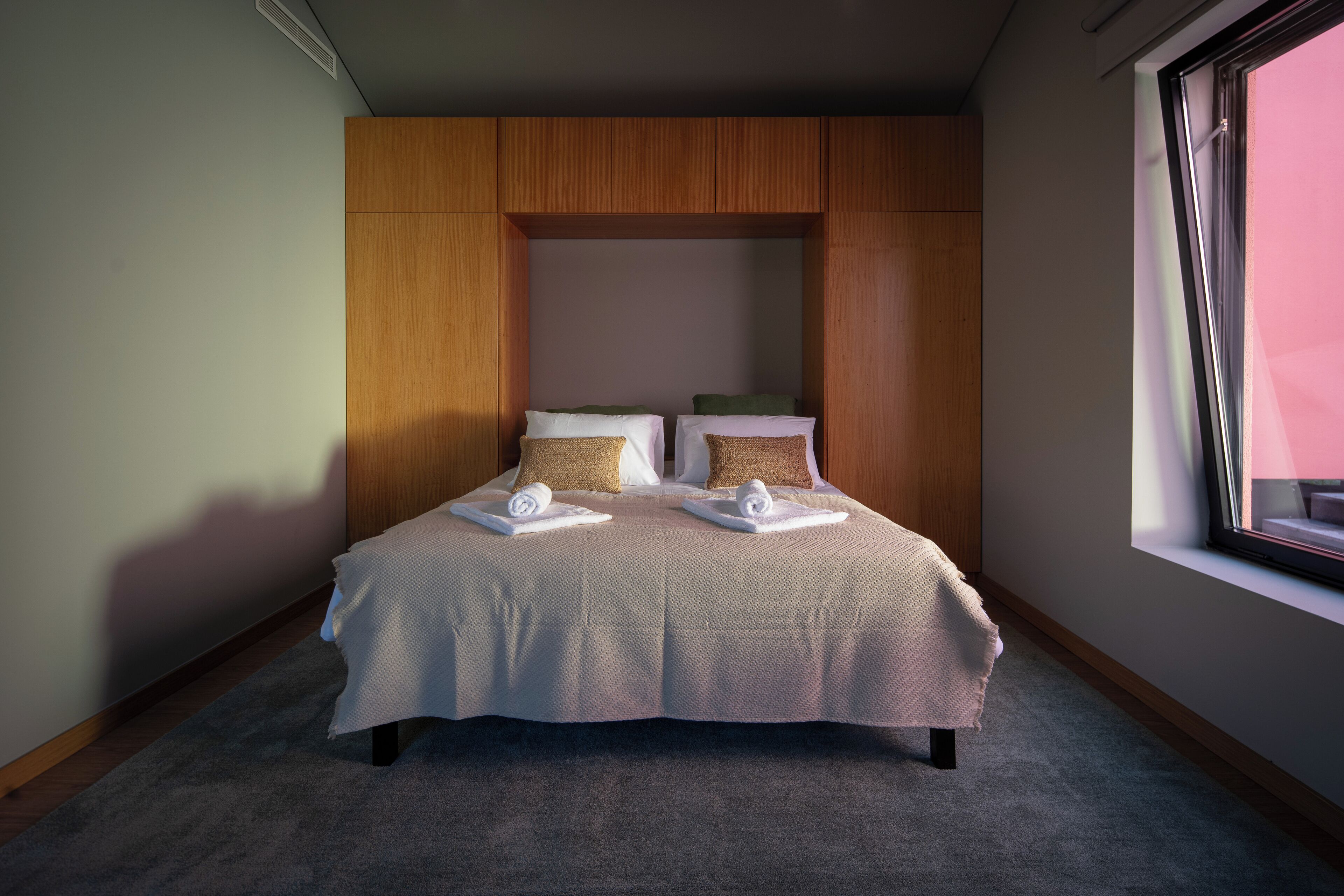 Ein ordentlich bezogenes Doppelbett mit einer beigen Tagesdecke, zwei Kissen und zwei aufgerollten Handtüchern. Der Raum verfügt über Holzvertäfelung an den Wänden und ein großes Fenster auf der rechten Seite.