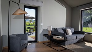 A modern living room with a grey sofa, a small armchair, a coffee table, and a floor lamp. There is a sliding glass door leading to an outdoor area with a table and chairs. - Canto's Green Garden & SPA  (Povoacao)