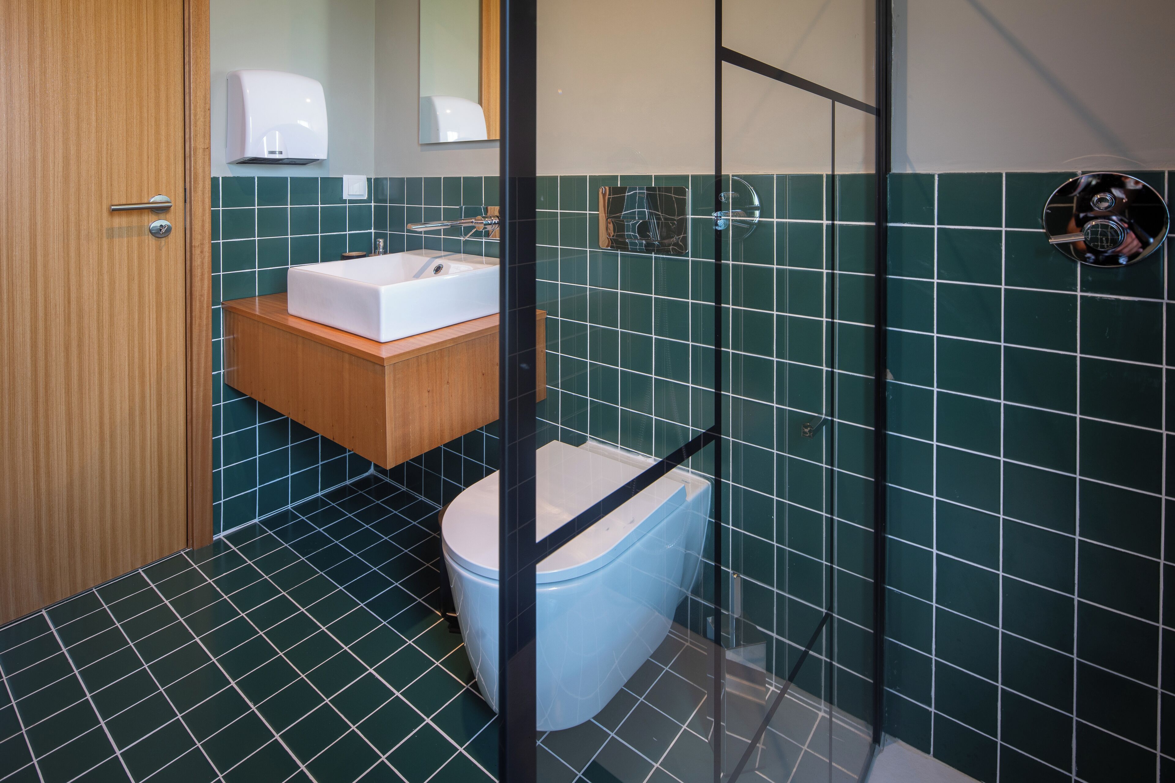 A bathroom with a wooden door, a white sink, a toilet, and green-tiled walls.