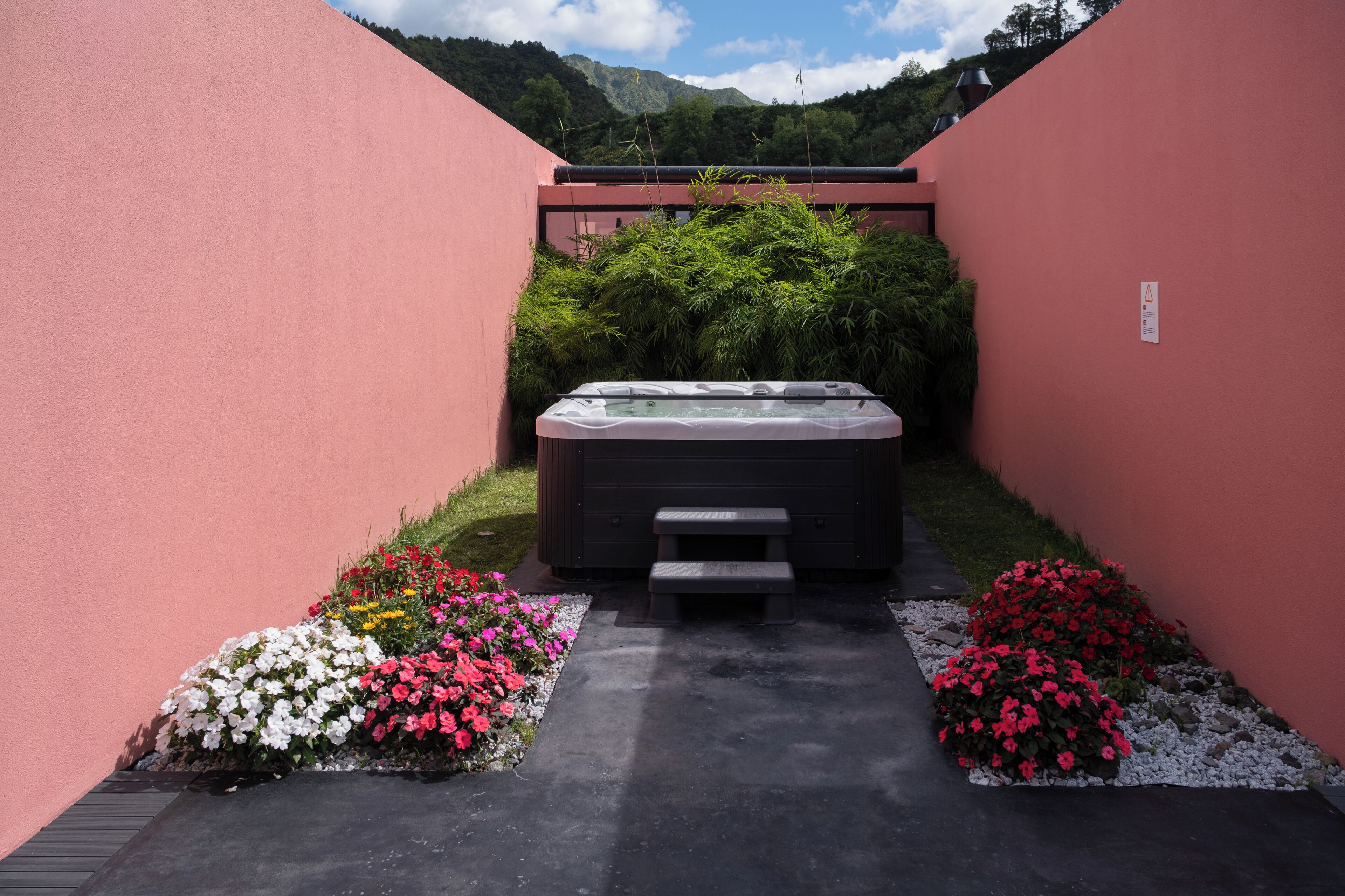 A hot tub in a narrow, pink-walled courtyard with flower beds on either side and a view of mountains in the background.