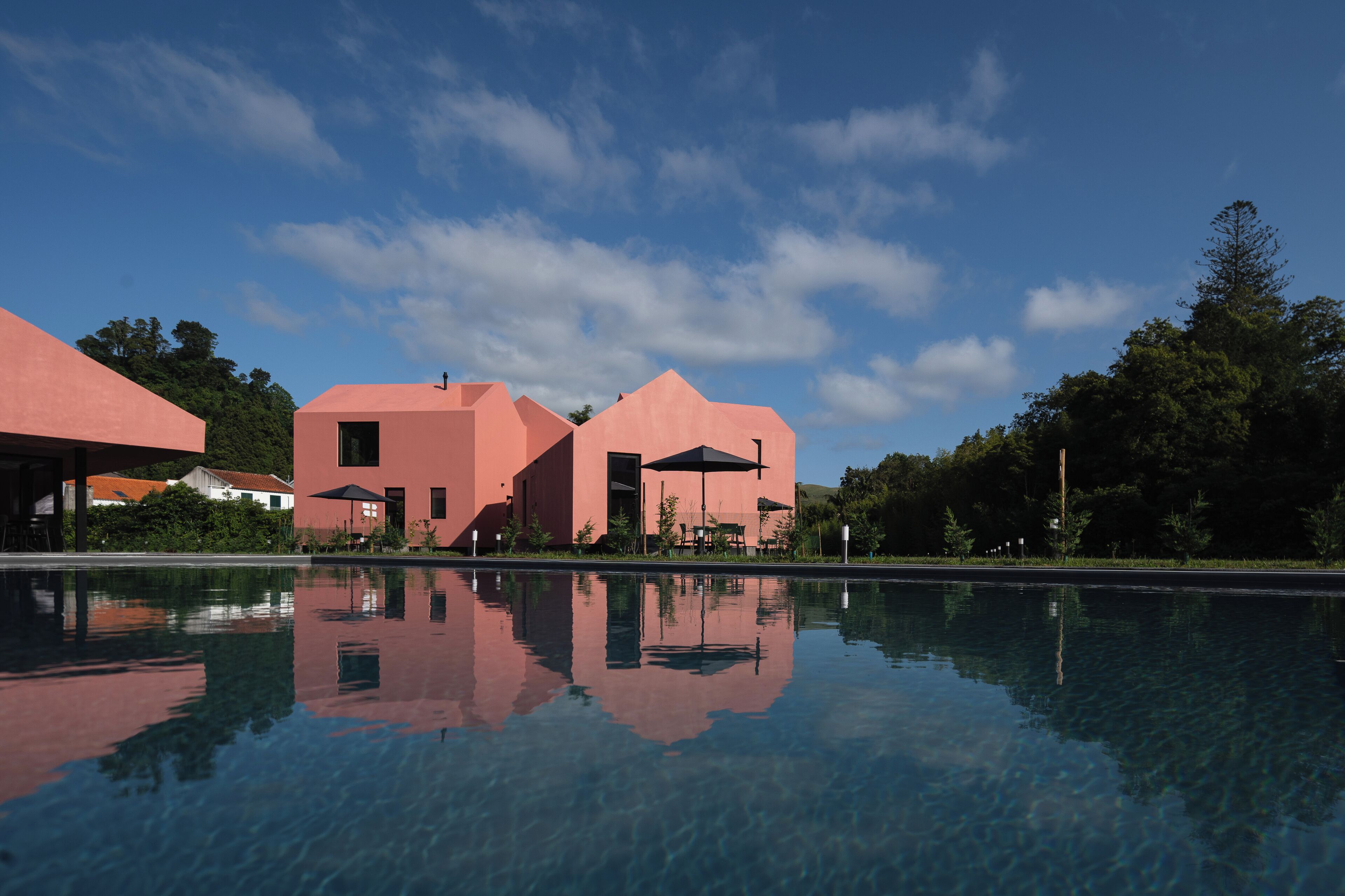 Un edificio moderno de fachada rosa, una piscina cristalina que refleja la estructura y follaje circundante.