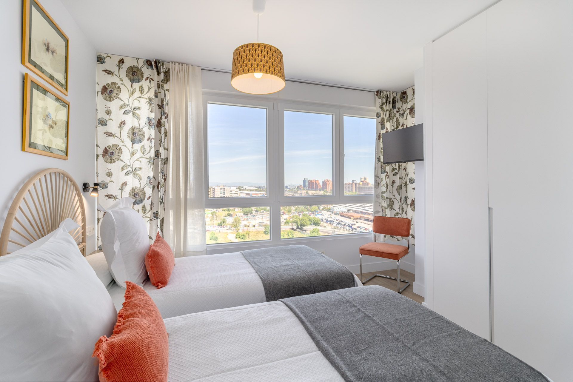 Ein Hotelzimmer mit zwei Betten, einem großen Fenster mit Blick auf die Stadt, einem an der Wand montierten Fernseher und einer gewebten Lampe.