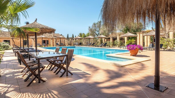 Ein Poolbereich mit Tisch und Stühlen, umgeben von Sonnenschirmen und Blumentöpfen.