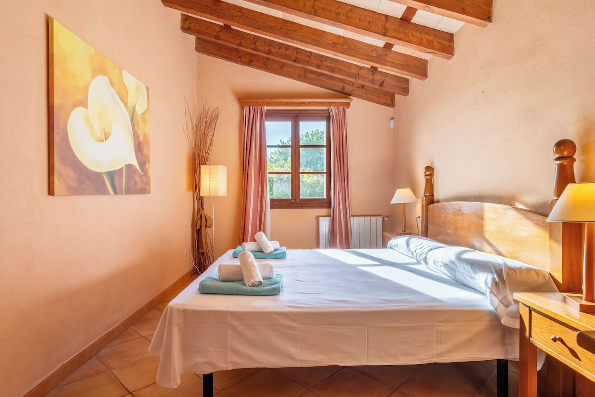 A bedroom with a bed, a window with curtains, a painting on the wall, and a lamp on the nightstand.