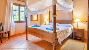 Ein Bett mit Himmelbett, ein Nachttisch mit Lampe, ein Sessel und ein Fenster mit Vorhängen.