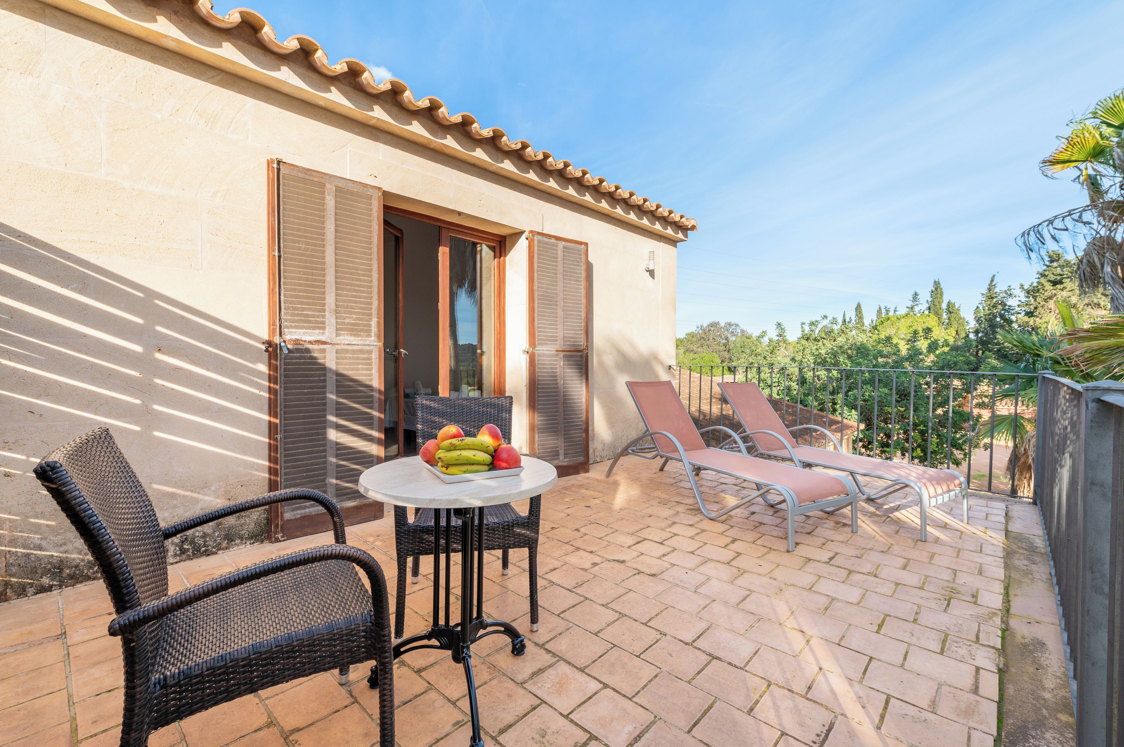 Une terrasse avec des chaises en osier, une table garnie de fruits et une vue sur des arbres et une clôture.