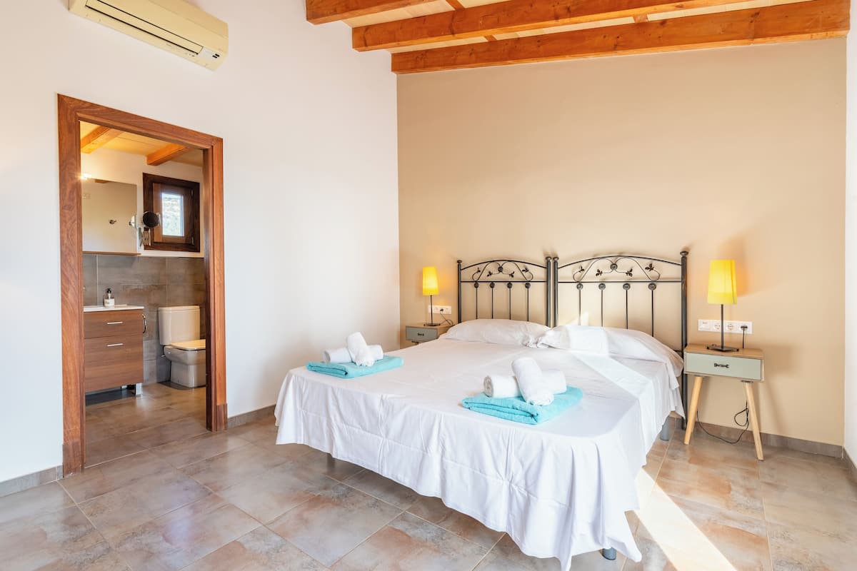 A bedroom with a bed, two bedside tables, a nightstand, a mirror, and a wooden door leading to a bathroom.