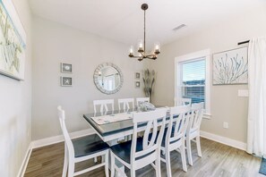 A dining area with a glass-top table, white chairs, a chandelier, and a window with blinds. - Modern 3BR, Dogs OK, Walk to beach, Hot Tub, Pool (Corolla)