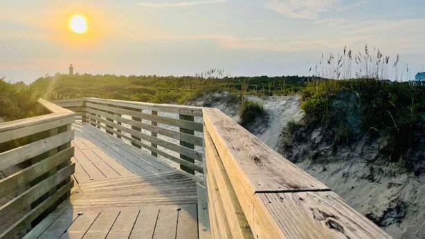 A wooden boardwalk leading to a dune landscape with a lighthouse in the distance.