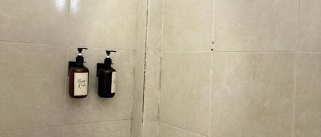 A shower area with beige tiles, a handheld showerhead, and two bottles of liquid soap on the wall.