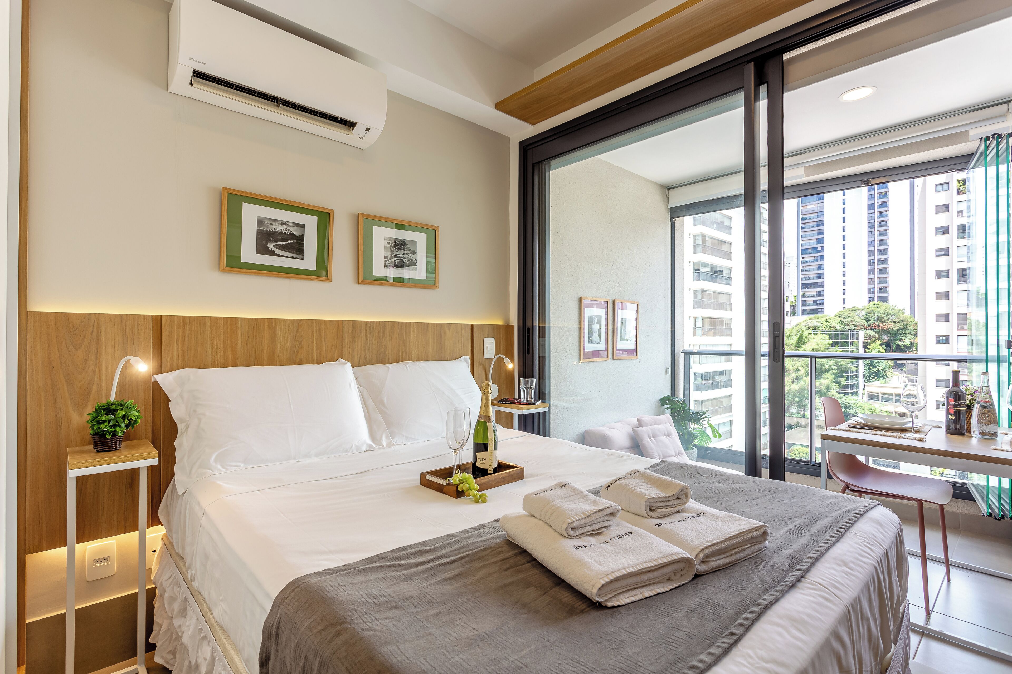 A modern hotel room with a large bed, a desk with a chair, and a balcony with a view of high-rise buildings.