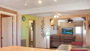 A living room with a large TV, seating area, and a ceiling fan. - Off The Hook (Port St. Joe)