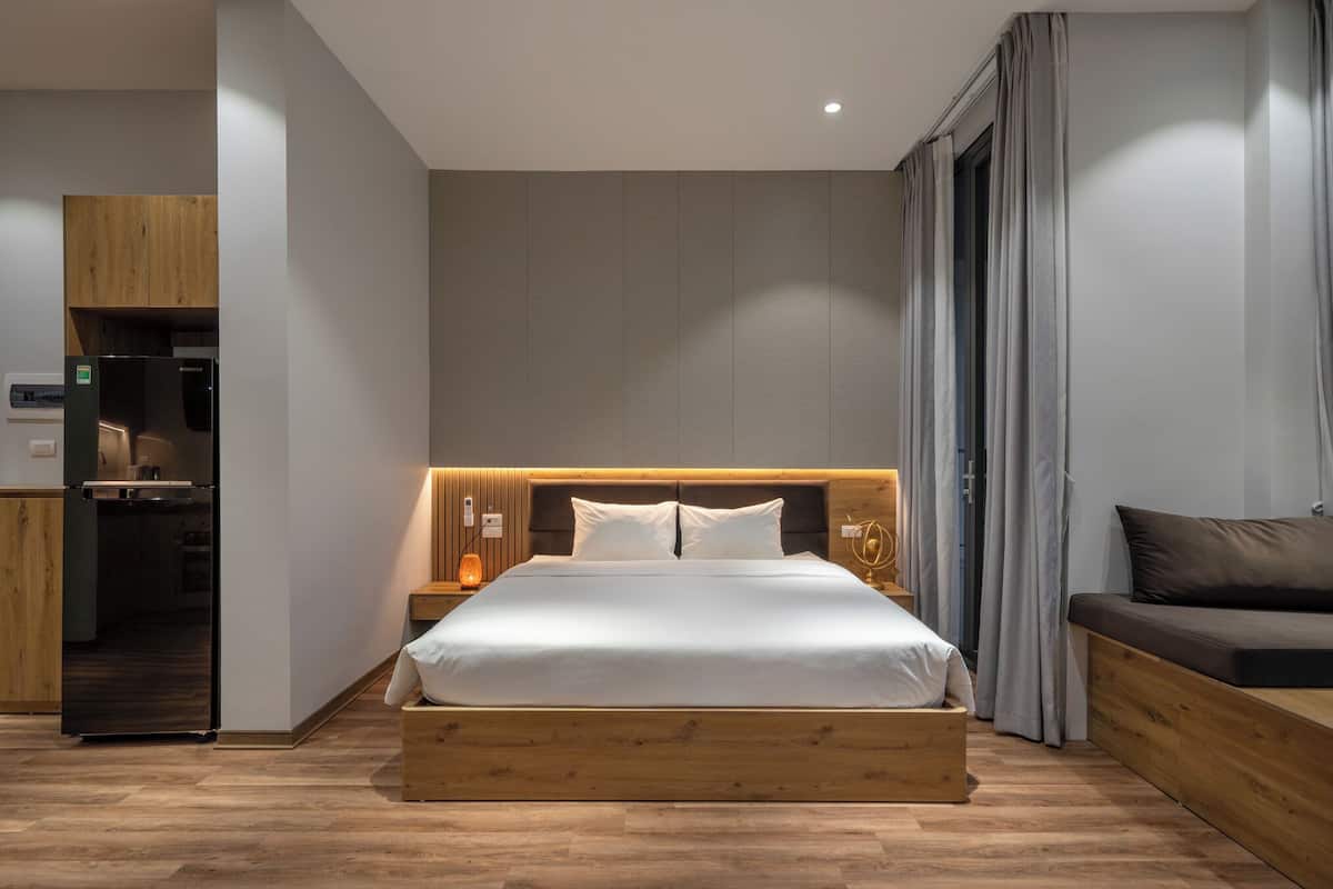 A modern bedroom with a wooden bed frame, a nightstand with a lamp, a built-in wardrobe, and a seating area with a sofa.