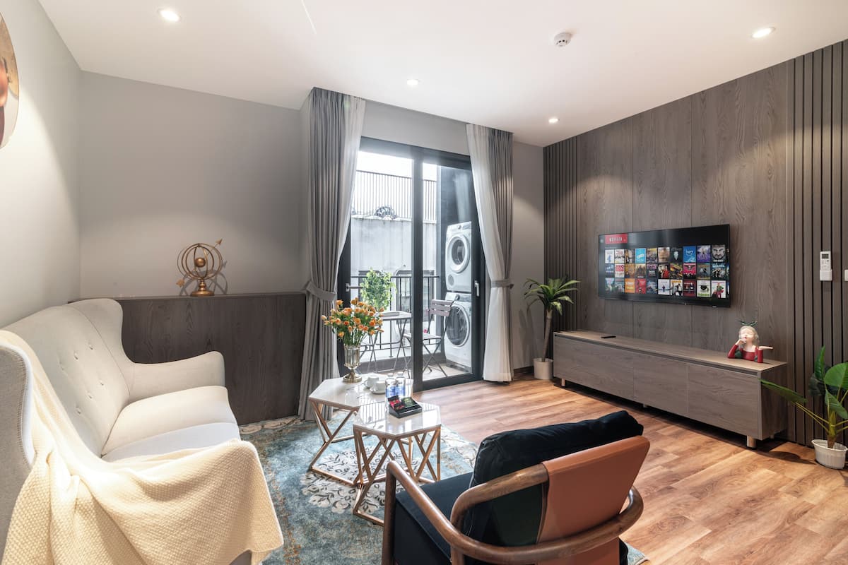A modern living room with a sofa, armchair, and coffee table, a flat-screen TV mounted on the wall, and a sliding glass door leading to a balcony with a washing machine.