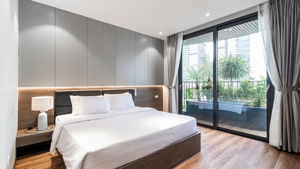 A modern hotel room with a large bed, a nightstand, a lamp, and a view of the outdoors through a sliding glass door. - Luxe Paradise Suites To Ngoc Van (Hanoi)
