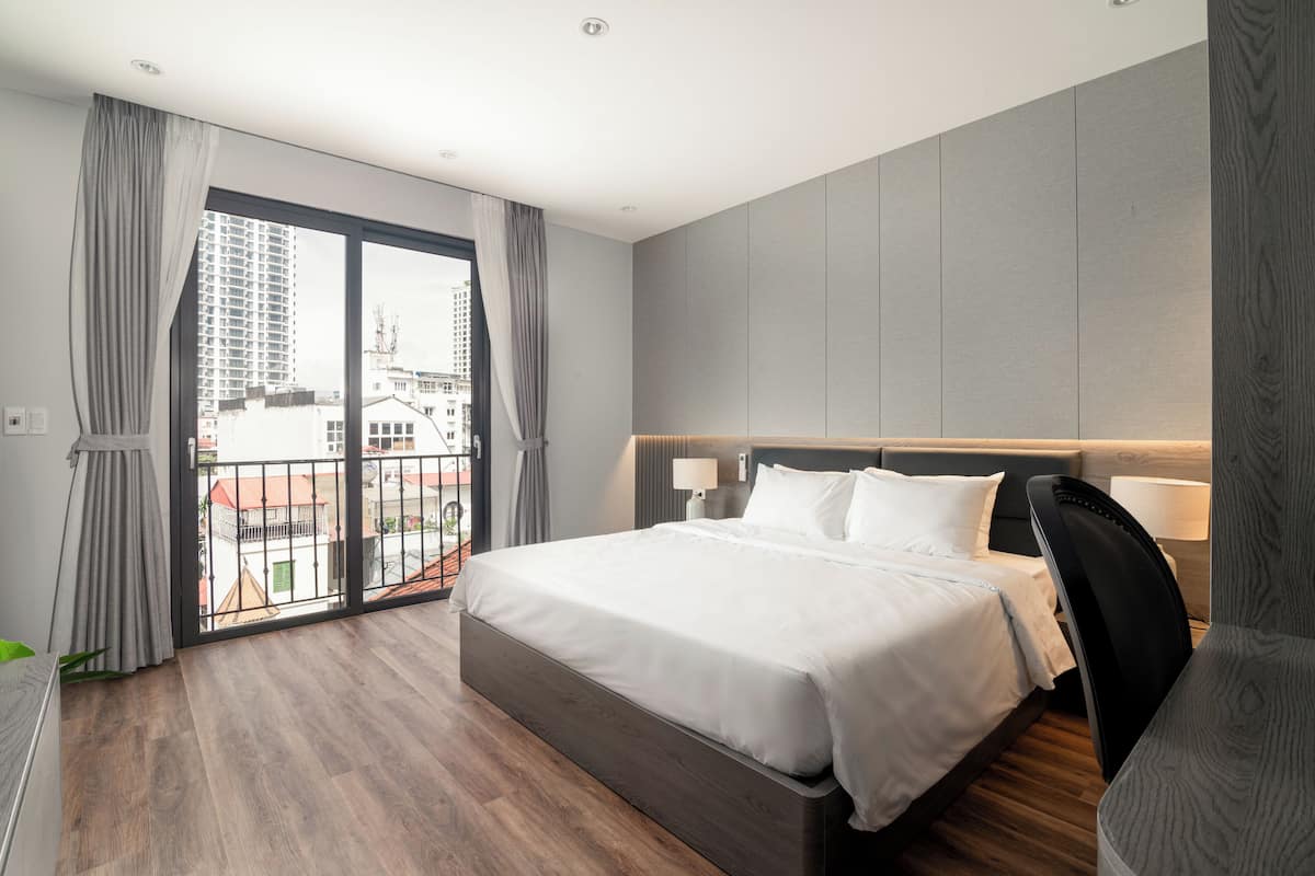 A modern hotel room with a large bed, a wooden floor, a balcony with curtains, and a city view.