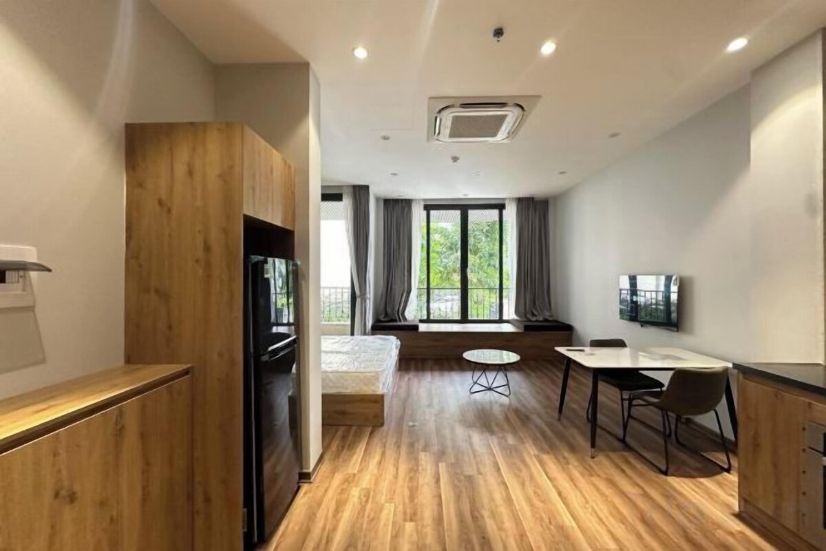Quarto de hotel moderno com piso de madeira, área de refeições com mesa e cadeiras, TV de tela plana na parede e uma grande janela com cortinas.