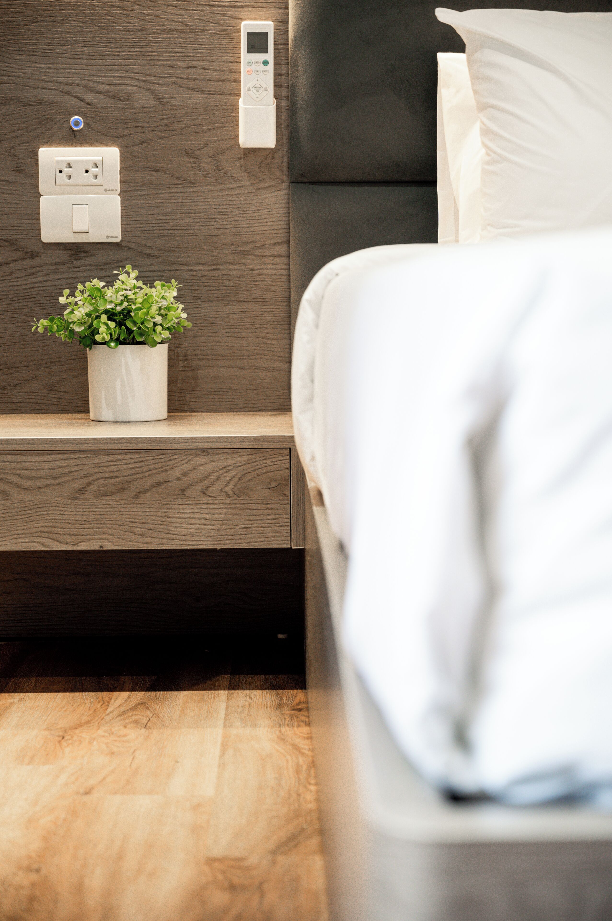Une chambre à coucher moderne avec une tête de lit en bois, une table de chevet ornée d’une plante en pot et un panneau de commande fixé au mur.