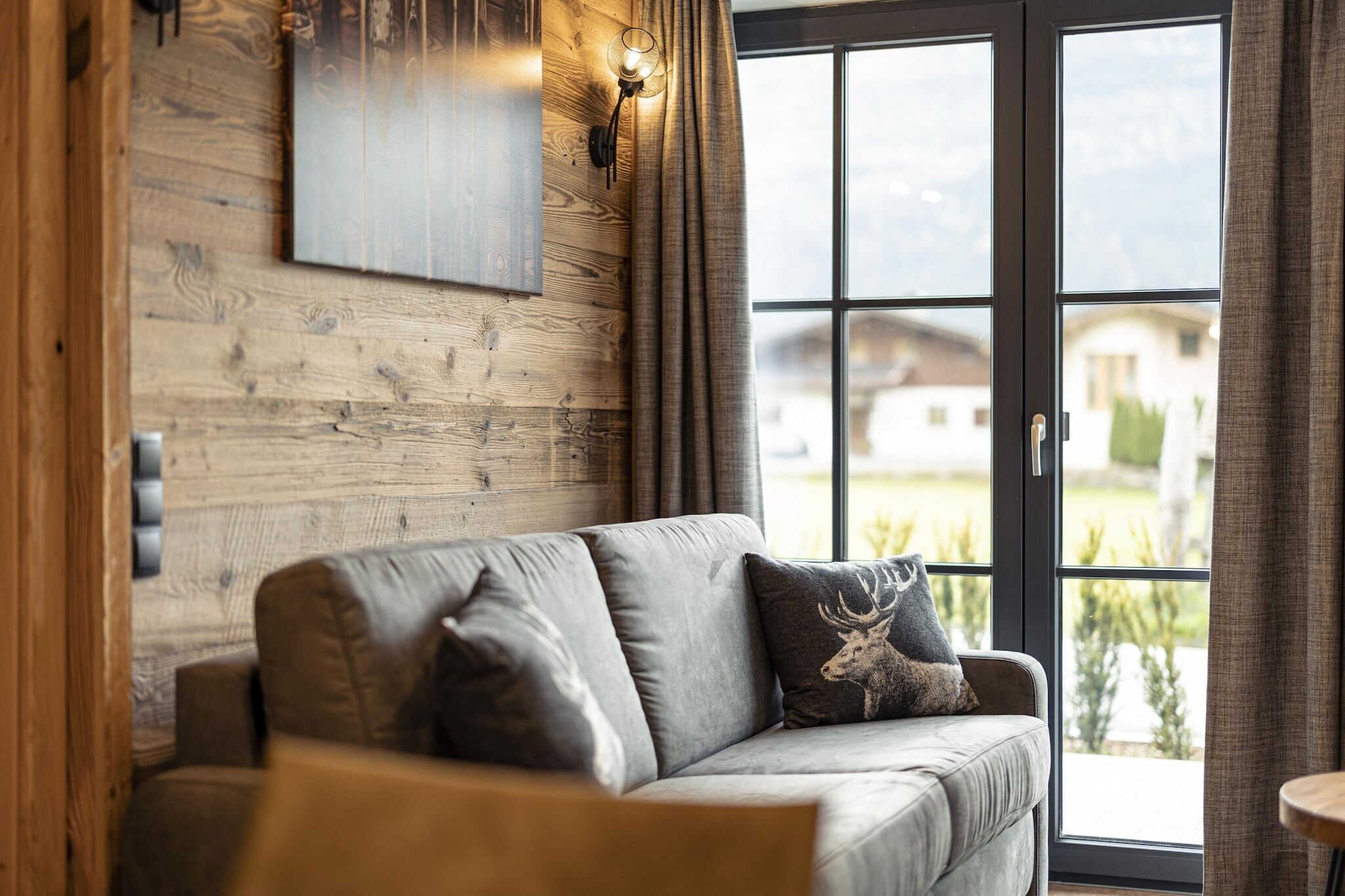 Ein gemütliches Wohnzimmer mit einer Holzwand, einem grauen Sofa mit Kissen mit Elchmotiv und einem großen Fenster mit Vorhängen.