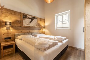 Un dormitorio con cabecera de madera, cama con sábanas blancas, lámpara de pared, ventana con persianas e imagen enmarcadas en la pared.