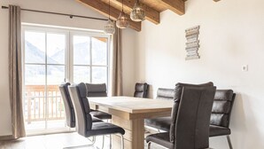 Un espace repas avec une table en bois et des chaises noires, de grandes fenêtres offrant une vue sur les montagnes et des lampes suspendues.