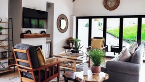 A modern living room with a wooden coffee table, a grey sofa, and a TV mounted on the wall. There is a large window with a view of greenery outside. - Indoni Eco Village  (Richmond)