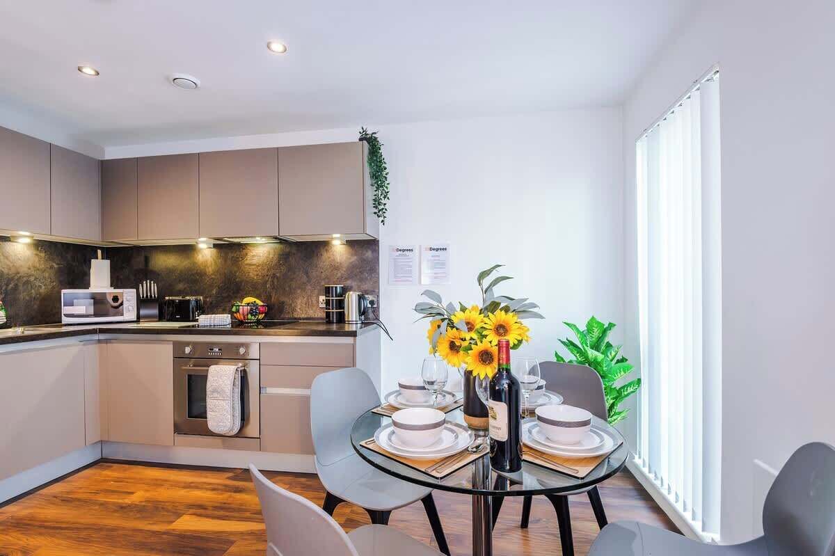 A modern kitchen with a dining area, featuring a glass table, chairs, and a vase with flowers.