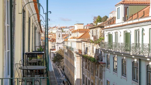 A narrow street with buildings on either side, featuring balconies with metal railings and a clock tower in the distance. - GuestReady - Night out adventure in Lisbon (Lisbon)
