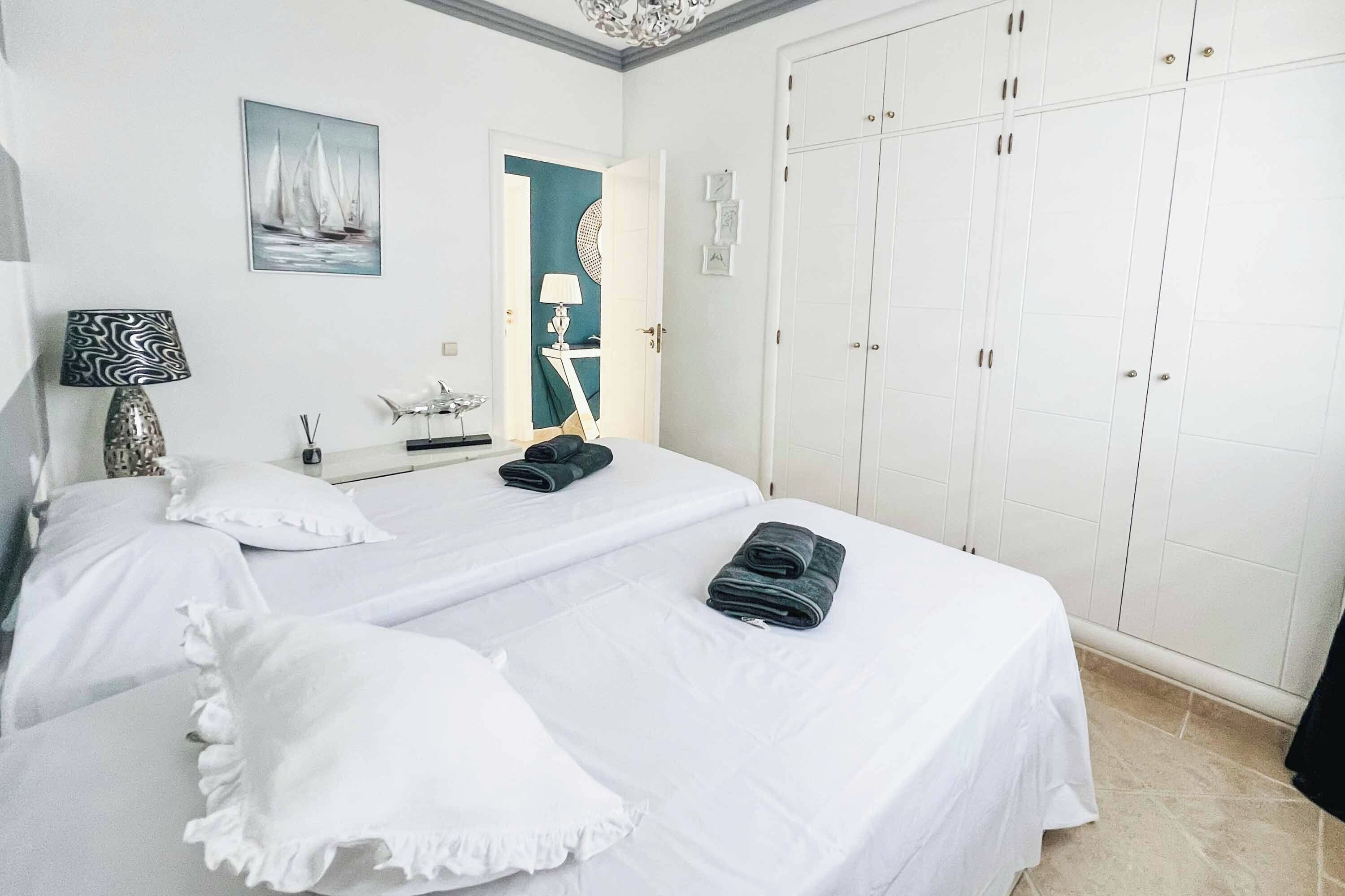 A hotel room with two beds, a large wardrobe, a painting of sailboats, a lamp, and a small table with a decorative item.