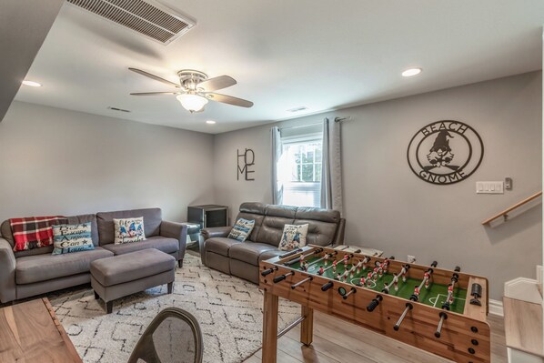 A living room with a foosball table, two sofas, a ceiling fan, and a wall with a beach-themed logo.