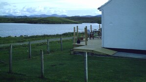 A lakeside view with a wooden deck and chairs overlooking a body of water, surrounded by grassy hills.