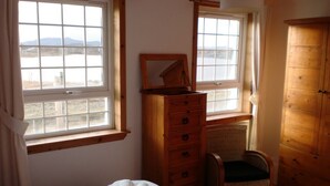 A bedroom with a bed, two windows, a wooden dresser, a chair, and a wardrobe.
