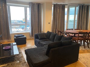 A living room with a sofa, ottoman, dining table, chairs, and a view of a cityscape through large windows. - London Riverside Apartment-2 Bed-freep-balcony (London)