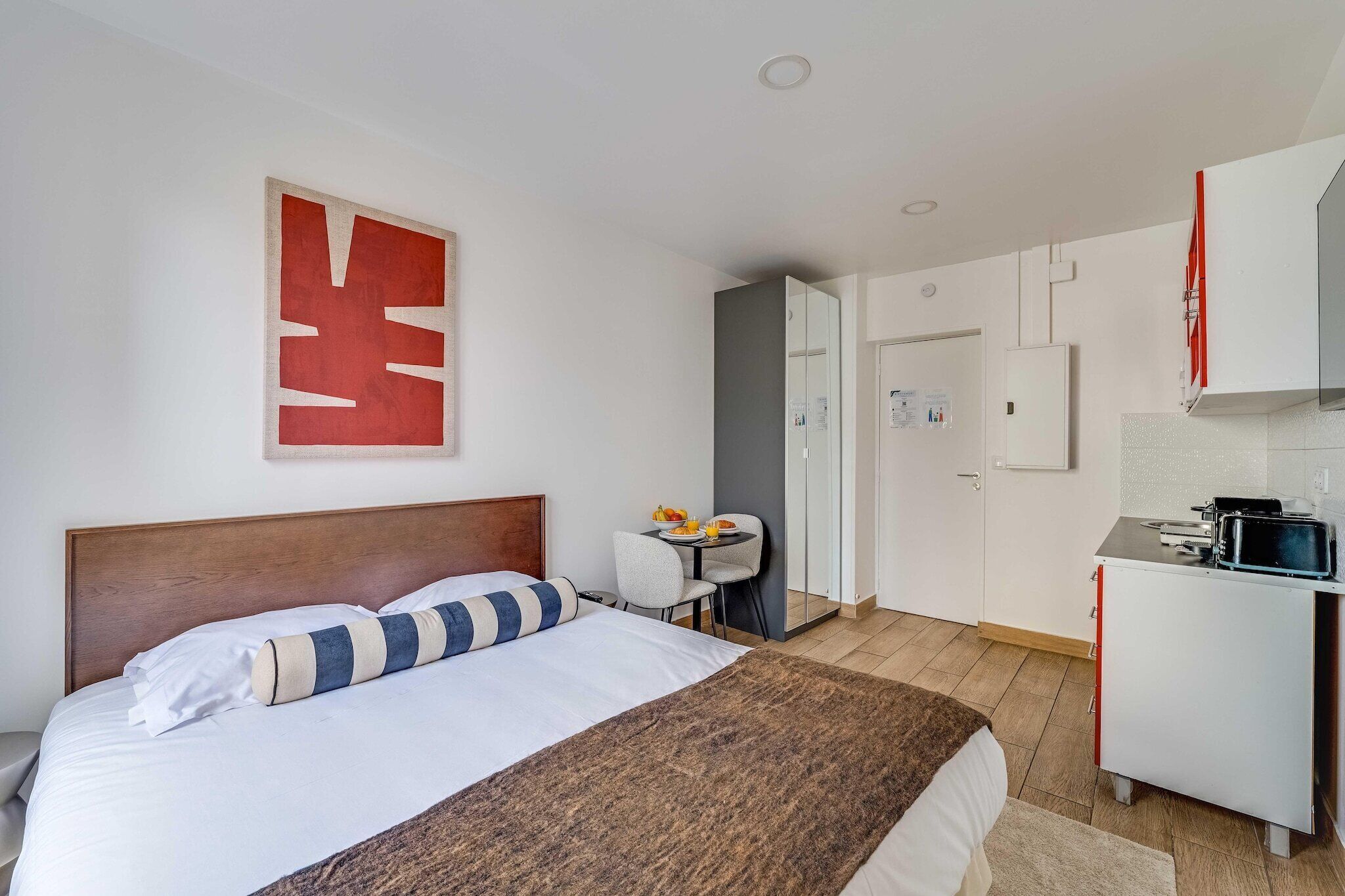 Quarto de hotel com cama, escrivaninha com cadeira, mesinha com prato e xícara, micro-ondas e geladeira.