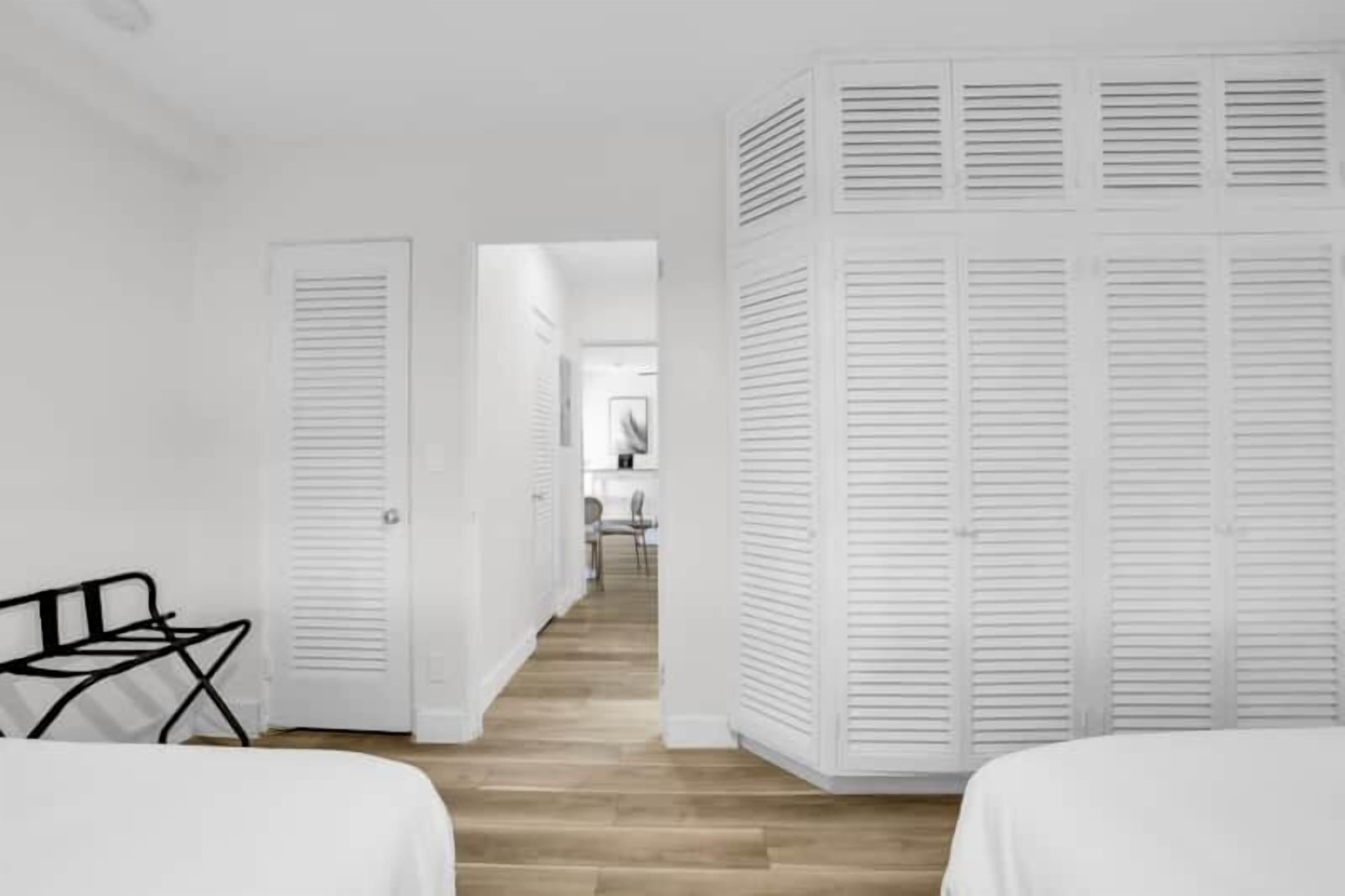 Habitación de hotel con piso de madera, paredes blancas y cama con ropa blanca. Hay una silla negra y un armario blanco con puertas persianas.