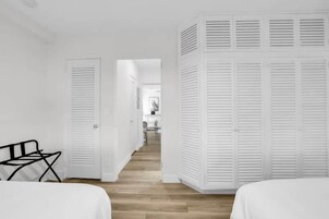 Habitación de hotel con piso de madera, paredes blancas y cama con ropa blanca. Hay una silla negra y un armario blanco con puertas persianas.