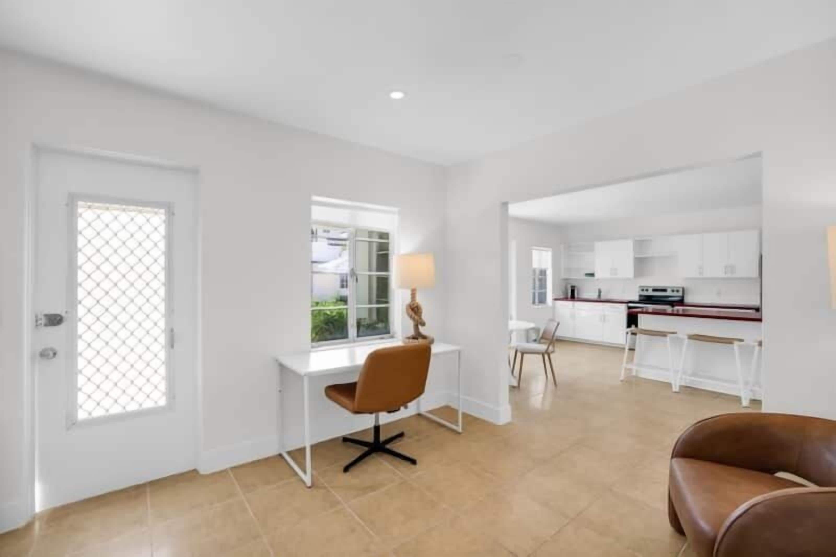 A spacious living area with a white desk, a brown chair, and a lamp. There is a kitchen area in the background with white cabinets and a red countertop.